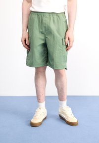 Pantalones cortos cargo verde claro hechos de un material ligero, con cinturilla elástica, bolsillos laterales y un corte holgado. Llevados con zapatillas beige.