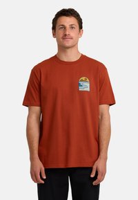 T-shirt z bawełny w kolorze rdzy z małym graficznym nadrukiem zachodu słońca i fal na lewej piersi. Krótkie rękawy, okrągły dekolt.