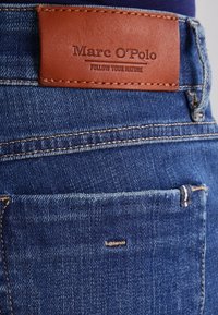 Jeans in denim blu con una toppa in pelle marrone con testo in rilievo. Cuciture visibili e un dettaglio a contrasto sulla tasca posteriore.