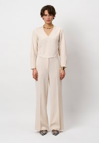 Beige jumpsuit met een V-hals, lange mouwen, een gladde voorkant en wijde pijpen. Gecombineerd met zwart-witte hakken met patroon. Gouden opvallende ketting.