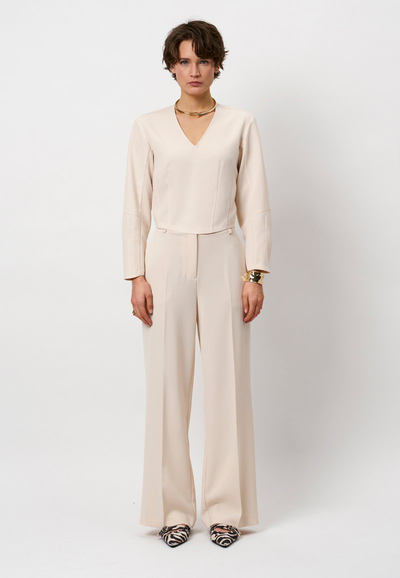 Beige jumpsuit met een V-hals, lange mouwen, een gladde voorkant en wijde pijpen. Gecombineerd met zwart-witte hakken met patroon. Gouden opvallende ketting.