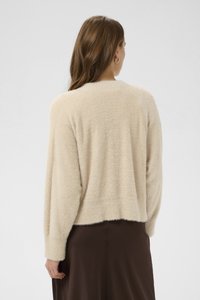 Žena s dlouhými hnědými vlasy, nosící zlaté kruhové náušnice, béžový chlupatý svetřík s dlouhým rukávem a hnědou sukni, otočená zády na bílém pozadí.