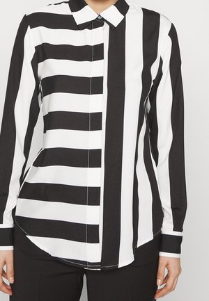 Mujer vistiendo una camisa abotonada a rayas blancas y negras, con rayas horizontales en el lado izquierdo y rayas verticales en el lado derecho.