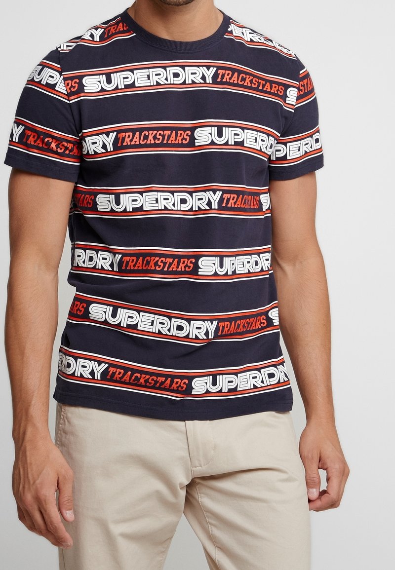 Marineblå bomulds T-shirt med gentagne hvide og orange "SUPERDRY TRACKSTARS"-tekster i vandrette striber. Rund hals, korte ærmer.
