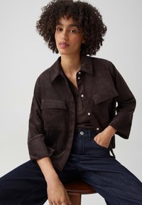 Young woman with curly hair sitting, wearing a dark brown corduroy jacket, matching top, and dark blue jeans against a plain background.