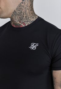 Melnā pieguļošā T-kreklā, kas izgatavots no gludas, elastīgas materiāla, kreisajā krūšu daļā ir baltā stilizētā logo. Īsas piedurknes.