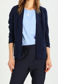 Marynarka w kolorze granatowym o luźnym kroju, z dwoma przednimi kieszeniami, w połączeniu z jasnoniebieskim t-shirtem zdobionym grafiką samolotu z papieru oraz spodniami w drobną prążki.