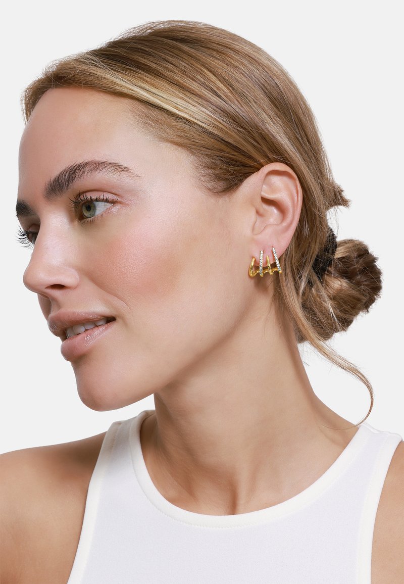 Boucles d'oreilles dorées avec trois anneaux décalés ornés de petites pierres claires, présentées sur un modèle aux cheveux châtain clair.