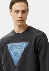 Felpa nera con colletto a coste, caratterizzata da un logo triangolare ricamato blu con il testo "GUESS U.S.A. WASHED JEANS".