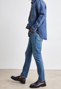 Tmavě modrá džínová košile s náprsní kapsou a zapínáním na knoflíky. Přiléhavé světle modré džíny a hnědé mokasíny s texturovanou podrážkou.