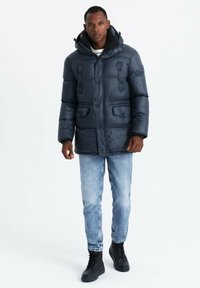 Giacca puffer blu navy con cappuccio, dotata di due tasche frontali, chiusure a scatto e una texture trapuntata. Abbinata a jeans azzurri e stivali neri.