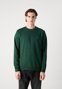 Felpa in cotone verde con girocollo, polsini e orlo a costine; presenta un piccolo logo sul petto sinistro; indossata con jeans scuri.