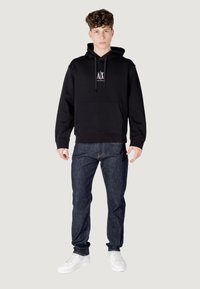 Felpa nera con cappuccio, con tasca frontale e cordino, abbinata a jeans blu scuro. Sneakers bianche completano l'outfit. Design semplice e moderno.