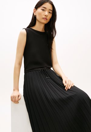 Jauna moteris su ilgais tamsiais plaukais, dėvinti juodą rankovių neturinčią palaidinę ir ilgą juodą klostuotą sijoną, sėdi ant balto bloko.