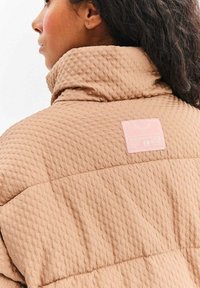 Béžová nadýchaná bunda s textúrovanou tkaninou a vysokým golierom. Na zadnej strane je ružová etiketa s pokynmi na údržbu.