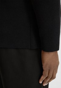 Czarny sweter dzianinowy o teksturowanej powierzchni. Posiada boczne rozcięcie i długie rękawy. Noszony na ciemnych spodniach, odsłaniający część ręki.