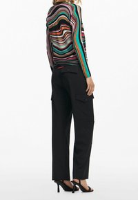 Femme portant un haut à manches longues rayé multicolore, un pantalon cargo noir avec poches à rabat, et des sandales noires à talons hauts, debout de profil.