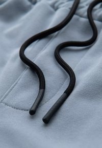 Tissu bleu clair avec une texture douce, présentant des cordons noirs fins avec des embouts en plastique, attachés à une taille élastique.