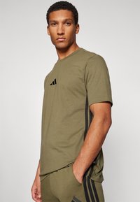 Olivkowo-zielony t-shirt sportowy z okrągłym dekoltem, z czarnymi paskami po bokach i małym czarnym logo na piersi. Materiał mieszany bawełniany.