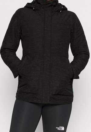 Person trägt eine schwarze Kapuzenjacke mit den Händen in den Taschen und schwarze North Face Leggings.