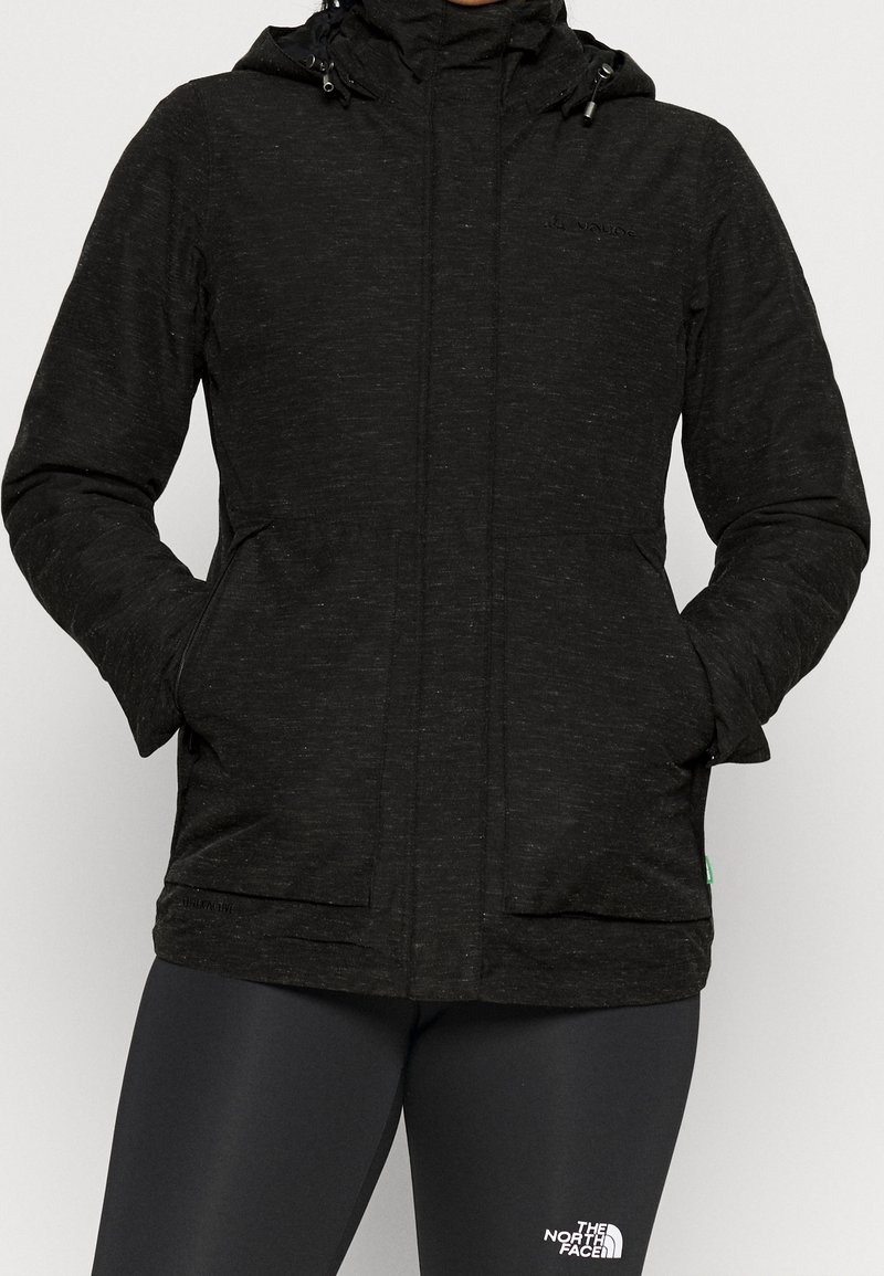 Personne portant une veste noire à capuche, les mains dans les poches, et des leggings noirs de marque North Face.