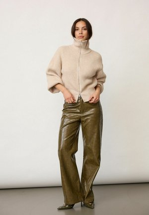 Woman wearing beige ribbed zip-up sweater and olive green wide-leg leather pants standing against plain background.