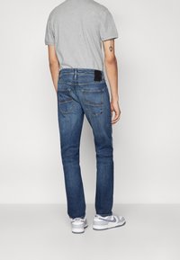 Jeans da uomo in denim blu con vestibilità dritta, due tasche posteriori e una targhetta in pelle nera sulla cinta, abbinati a scarpe sportive bianche.