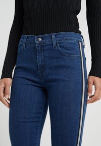 Skinny jeans in dark blue denim, featuring a mid-rise waist, five-pocket styling, and black and white side stripes along the legs.