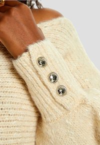 Maglione lavorato a maglia in crema con polsini a coste e tre bottoni argentati. La texture è morbida e pelosa, evidenziando il suo design caldo.