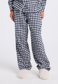 Marineblauwe en witte geblokte pyjamabroek gecombineerd met een bijpassend bovenstuk en fuzzy crème gekleurde slippers op een staande persoon.