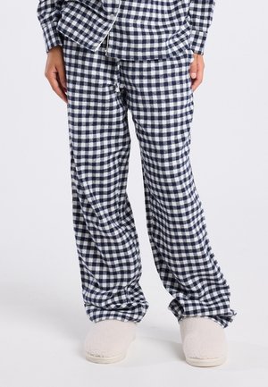 Un pantalon de pyjama à carreaux bleu marine et blanc associé à un haut assorti et des pantoufles crémeuses duveteuses sur une personne debout.