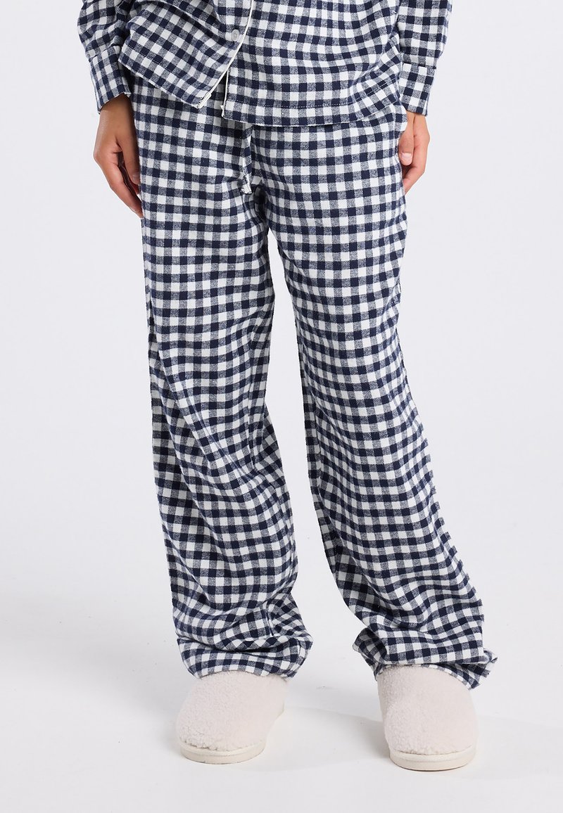 Marineblauwe en witte geblokte pyjamabroek gecombineerd met een bijpassend bovenstuk en fuzzy crème gekleurde slippers op een staande persoon.