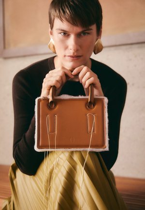 Sac en cuir brun avec une doublure blanche douce, présentant deux poignées courtes et des coutures visibles. La modèle tient le sac contre une jupe jaune plissée.