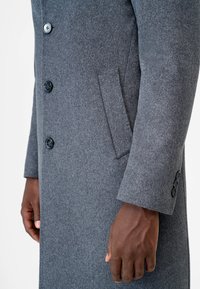 Grijze wollen jas met een getailleerde pasvorm, voorzien van grote knopen, een zijzak en een gemêleerde stof. Detail omvat een manchet met drie knopen.