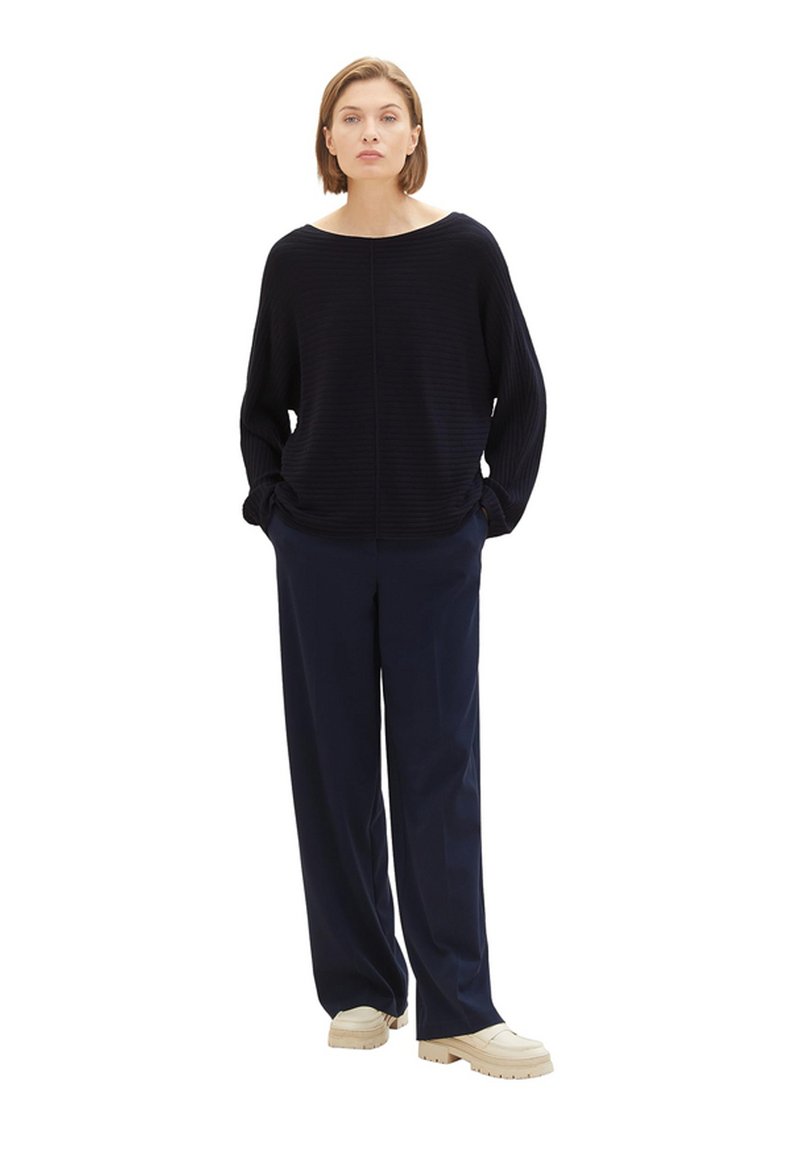 Marineblaues Rippstrickpullover mit weitem Halsausschnitt, überschnittenen Schultern und langen Ärmeln, kombiniert mit weiten marineblauen Hosen und hellen Sneakers.