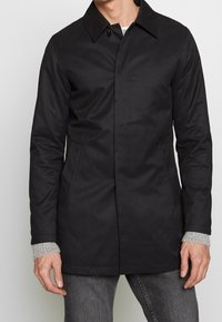 Muž v černém zapínacím kabátu přes šedý svetr a tmavé džíny, zobrazený od krku po střed stehenní oblasti, stojící na pozadí bez vzoru.