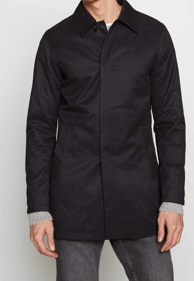 Muž v černém zapínacím kabátu přes šedý svetr a tmavé džíny, zobrazený od krku po střed stehenní oblasti, stojící na pozadí bez vzoru.