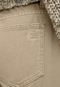 Poche en tissu beige d'un pantalon avec des détails de couture. Un logo embossé est visible sur la partie supérieure de la poche.