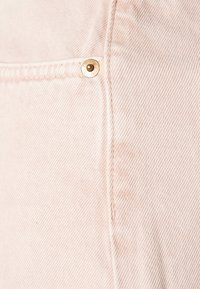 Vaaleanpunainen farkkukangas, jossa on vinoraidallinen rakenne ja kullanvärinen niitti sekä näkyvät ompeleet taskun sauman kohdalla.