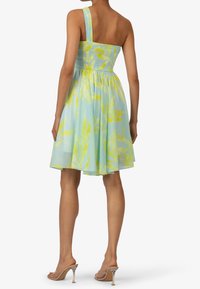 Robe bleu clair avec motif de feuilles jaunes, une bretelle, bodice smocké, jupe évasée et talons transparents portés avec la robe.