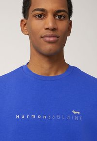 Blauwe katoenen crewneck t-shirt met een wit logo "Harmont & Blaine" en een geïllustreerde teckelaccent aan de linkerkant.