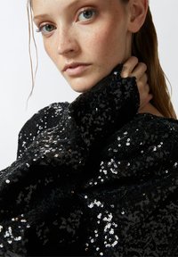 Crna bluzica sa šljokicama visokog dekoltea i teksturiranim rukavima, sa crnim dugmadi na manžetama. Sjajna, reflektirajuća površina.