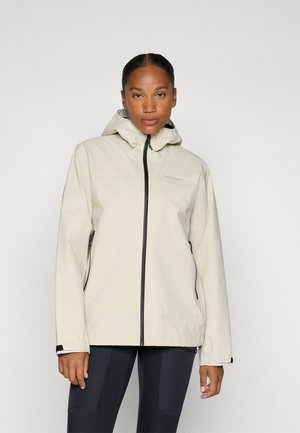 Femme portant une veste à capuche beige clair avec une fermeture éclair noire et un pantalon foncé, debout devant un fond blanc uni.