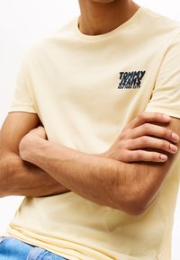 Ljusgul bomullströja med korta ärmar och rund halsringning. Har en marinblå grafisk tryck med texten "TOMMY JEANS NEW YORK CITY."