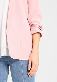 En kvinnas arm och hand syns, iklädd en ljusrosa blazer med uppvikta ärmar, vit blus och blå denimjeans mot en enfärgad bakgrund.