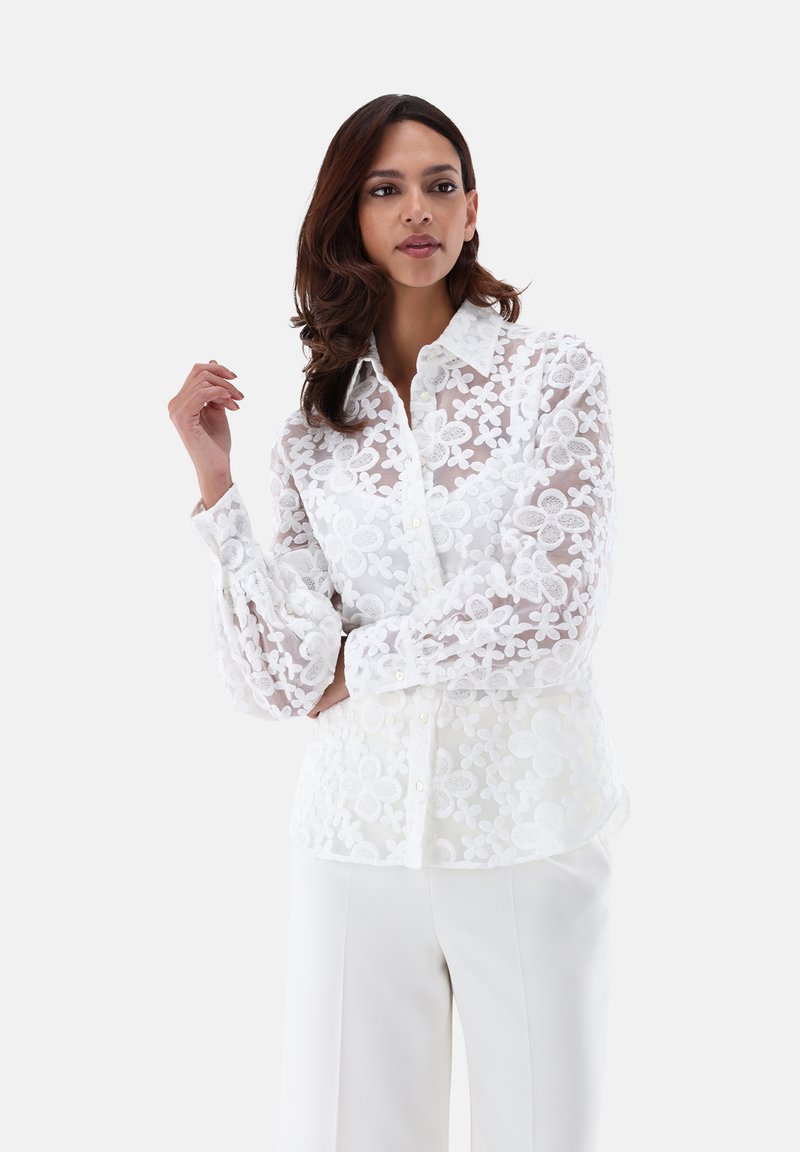 White floral lace shirt with long sleeves, collared neckline, and button closure. Features sheer fabric with textured flower patterns.