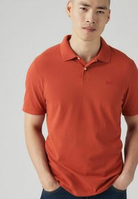 Kortärmad polo skjorta i livlig orange, gjord av bomull, med krage, treknapplist och en liten logotyp på vänster bröst.