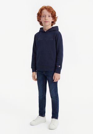 Barn med rødt hår iført navy Champion hættetrøje, mørke jeans og hvide sneakers, der står mod en hvid baggrund.