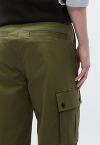 Olivzaļas kravas bikses ar gludu tekstūru, ar sānu kabatām ar piespraužamām pogām un aizmugurējām aplikācijas kabatām.