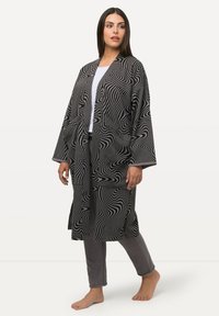 Schwarz-weiß gemusterter Morgenmantel mit langen Ärmeln und offener Vorderseite. Weicher Stoff mit wellenförmigem Design, kombiniert mit grauen Hosen und barfuß.